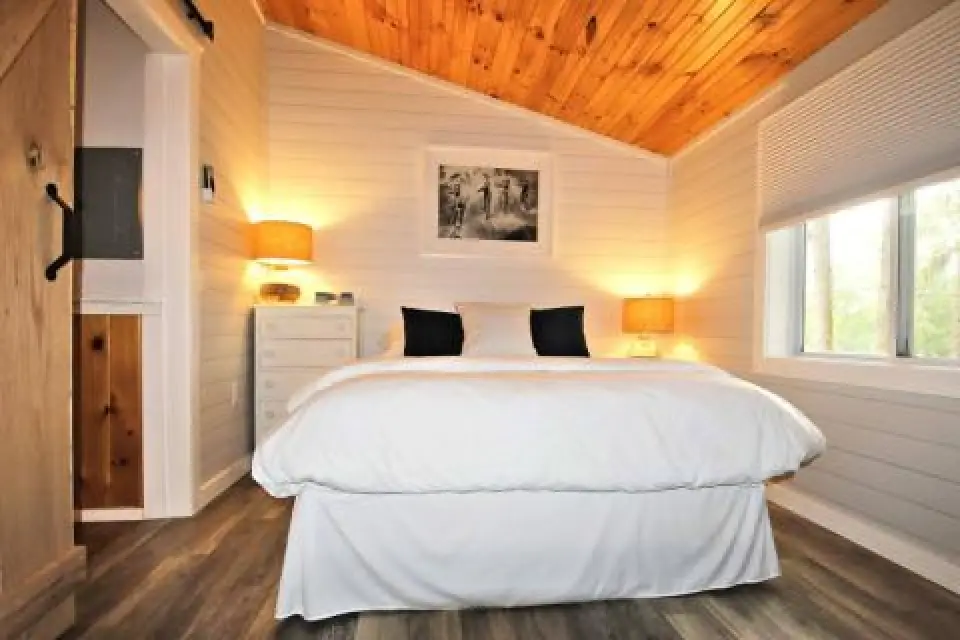 Cozy bedroom with white bedding, wood ceiling, two lamps, and window letting in natural light.