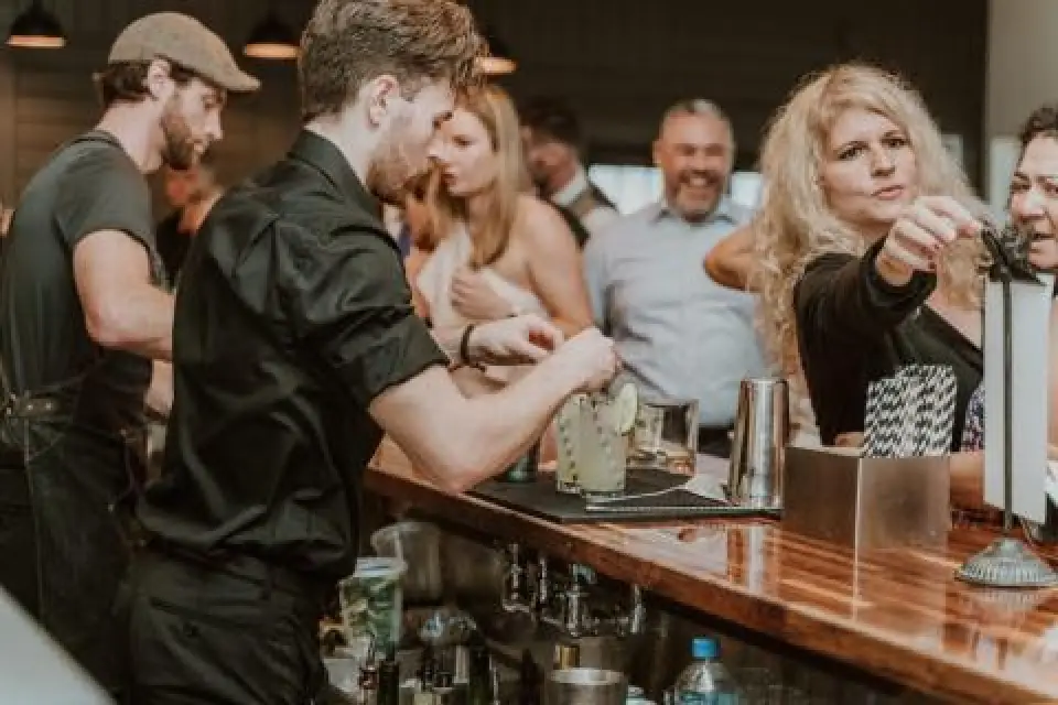 A bartender prepares drinks as people socialize and smile at a busy bar.