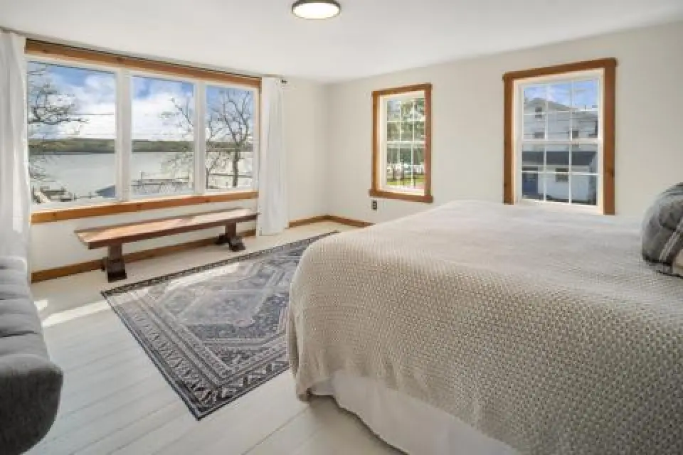 Bright bedroom with large windows, bed, patterned rug, and bench overlooking a lake view.