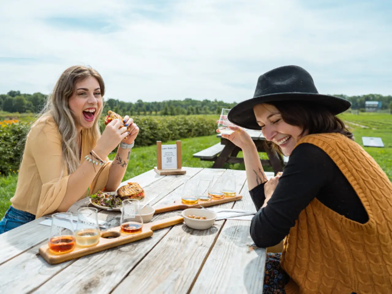 Two women enjoying food and drinks at an outdoor picnic table in a sunny field.