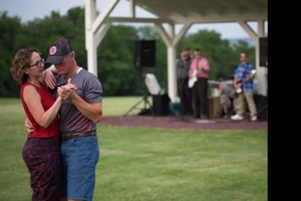 A couple dances outdoors on grass while a band performs under a pavilion in the background.
