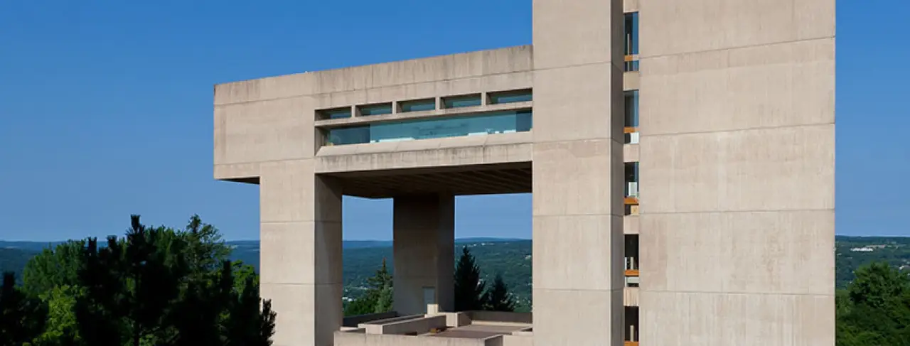 A modernist concrete building with a skywalk, set against a blue sky and green landscape.