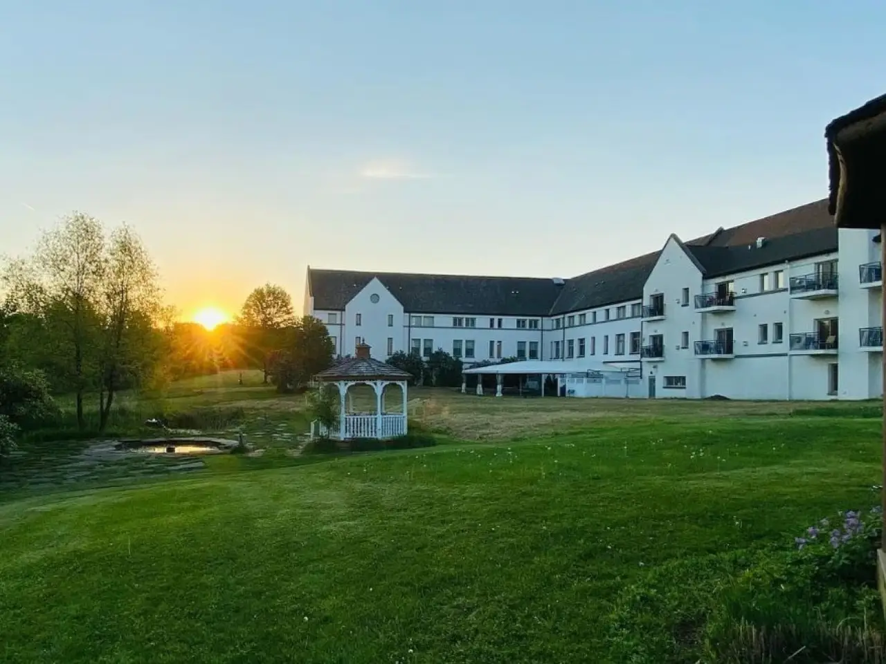 Sunset behind trees near a large white building with balconies and a gazebo on green lawn.