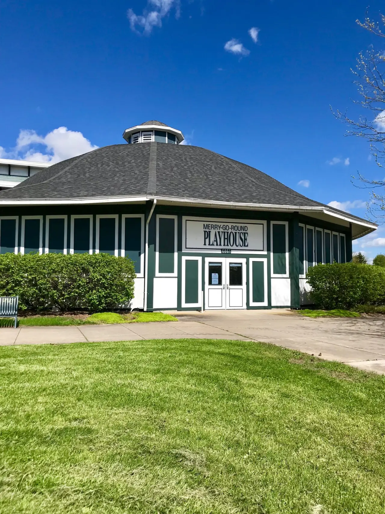 Octagonal building with green and white walls labeled “Merry-Go-Round Playhouse” under blue sky.