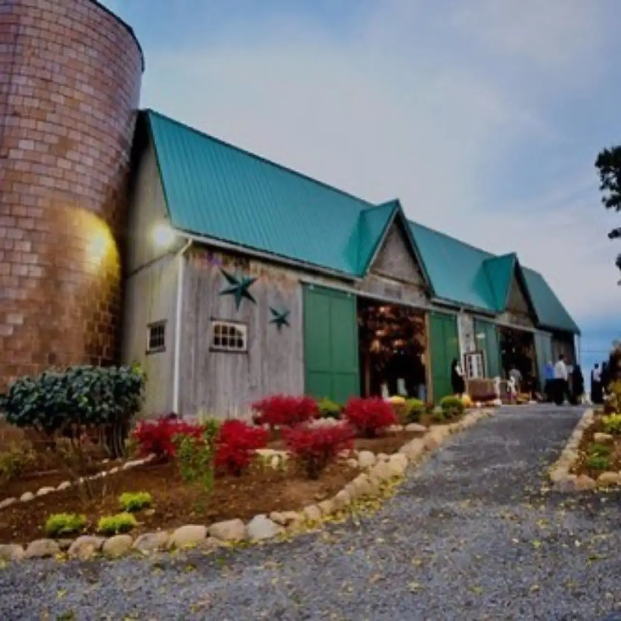 A barn with a green roof, silo, and garden bed lined with rocks and red bushes.