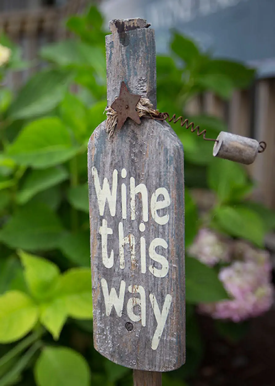 rustic sign shaped like a bottle reads Wine this way with greenery in the background