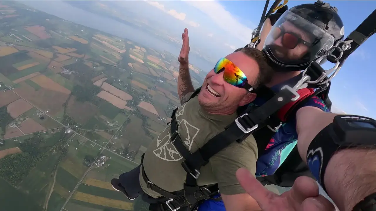 Two people tandem skydiving high above patchwork fields, one smiling with sunglasses on.