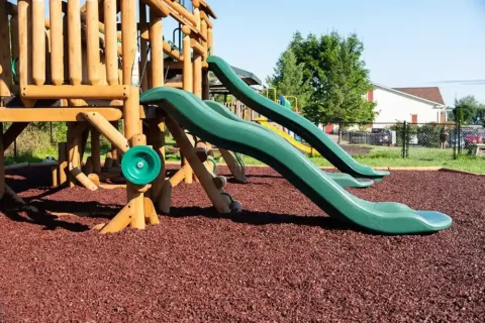 Two green slides on a wooden playground set over brown mulch, with trees and houses nearby.