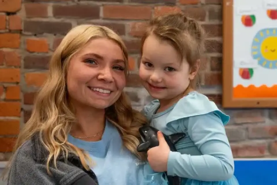 A smiling woman holds a young girl in a blue dress in front of a brick wall.