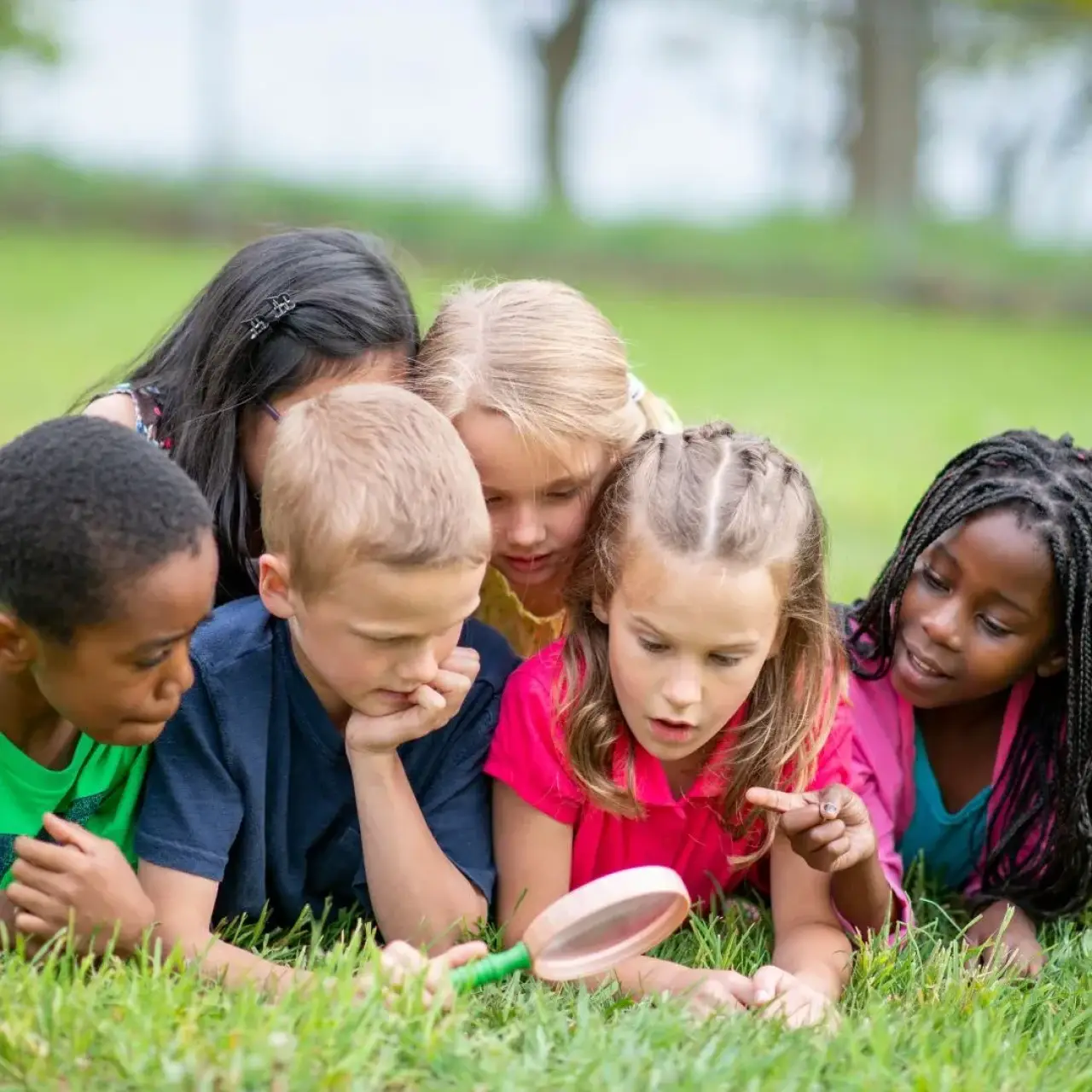 Six children lie on grass, looking closely at something with a magnifying glass.