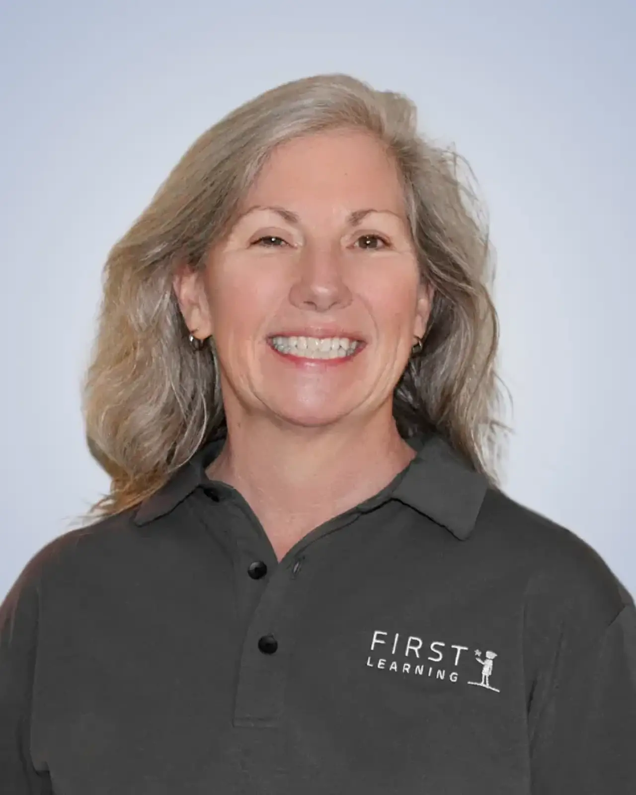 Smiling woman with gray hair wearing a dark polo shirt that says First Learning on it.