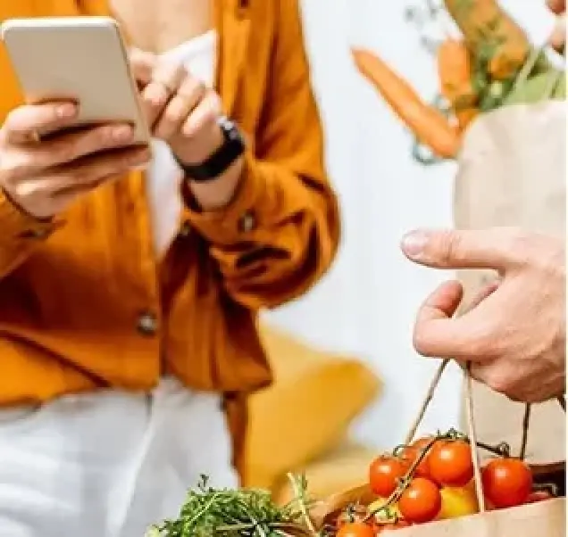 woman in orange blouse showing another person their phone while holding groceries