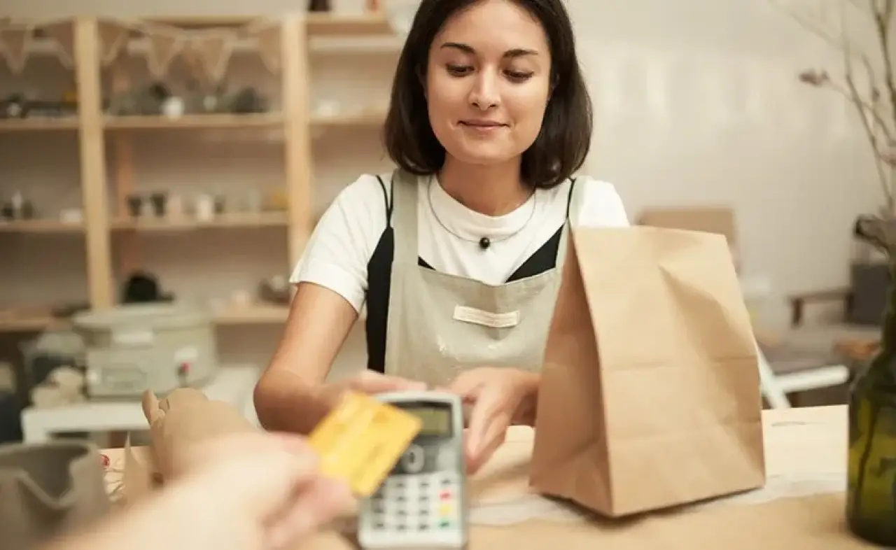 worker in white shirt and tan apron holds out payment processor to a customer