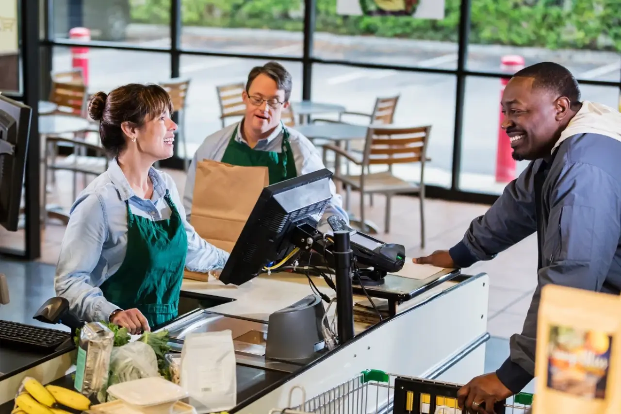 Cashier and bagger assist a smiling customer at a grocery store checkout.