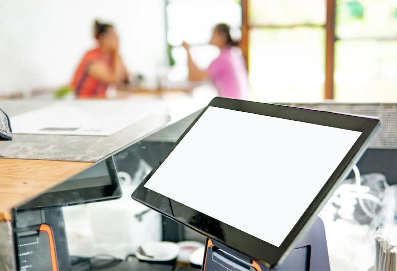 touchscreen pos system in foreground with two customers chatting at a table in background