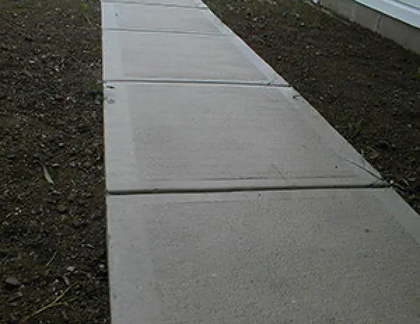 Concrete sidewalk slabs bordered by bare soil, leading alongside a building.