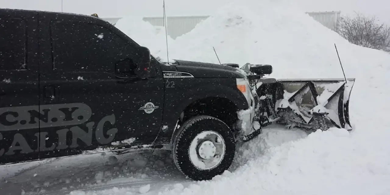A black pickup truck with a snowplow clears deep snow in a snowy, winter setting.