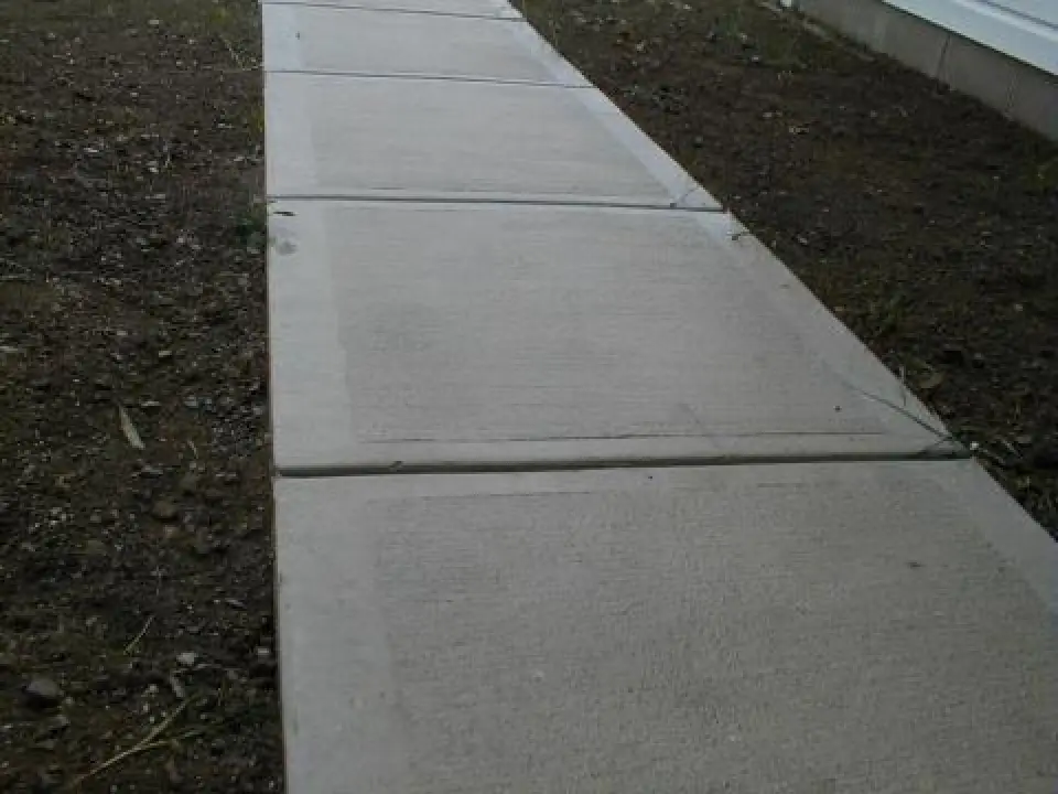A concrete sidewalk with square sections runs beside bare soil and a building wall.