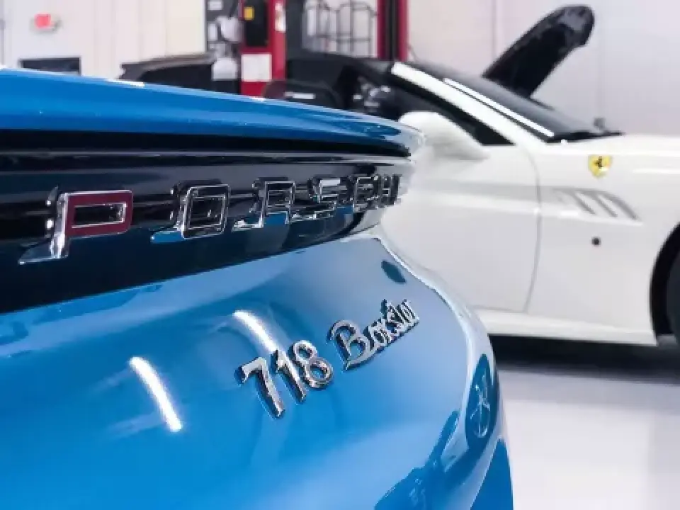 Close-up of a blue Porsche 718 Boxster badge, with a white Ferrari in the background.