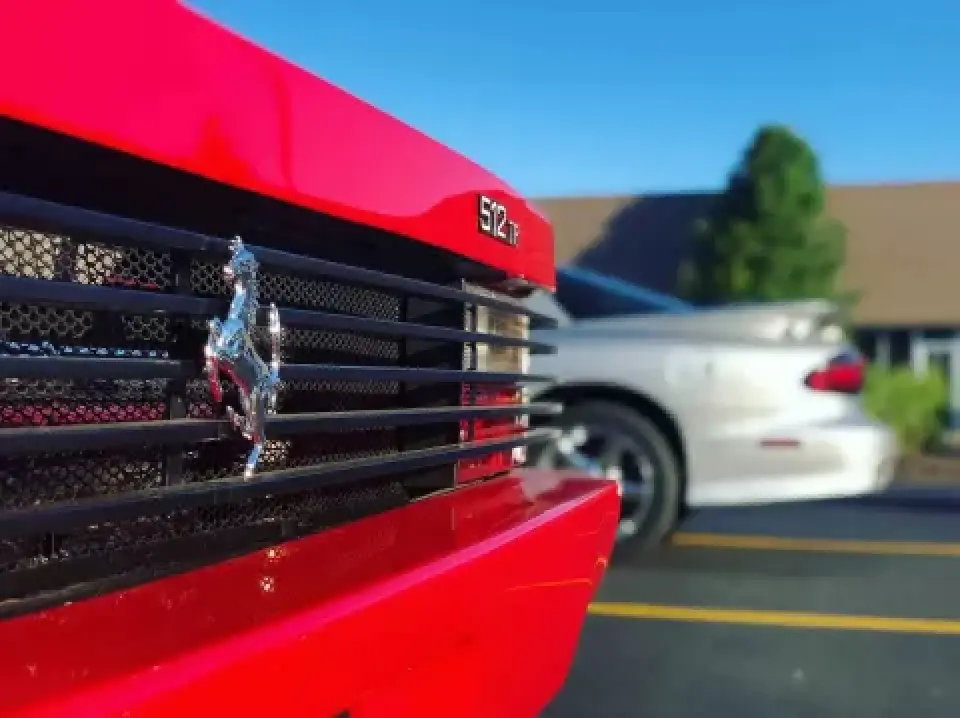 Close-up of a red Ferrari 512 TR grille, with a silver car blurred in the background.