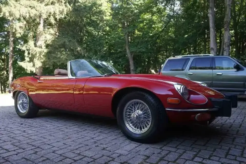 A red vintage convertible sports car parked on a paved area near a green SUV and trees.