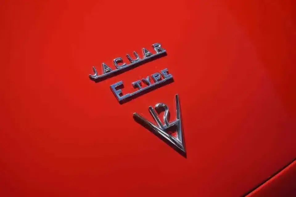 Chrome Jaguar E-Type V12 badge on a shiny red car hood.