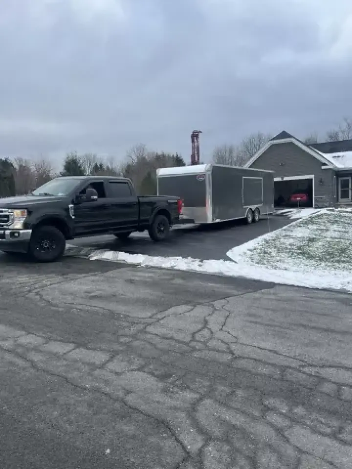 A black pickup truck towing a large silver trailer on a snowy driveway near a house.