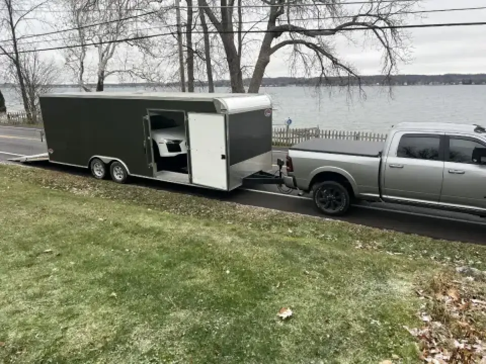 A silver pickup truck towing a large enclosed trailer with a car inside, parked by a lake.