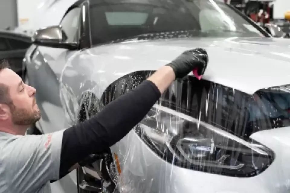 Person applying protective film to the front of a silver car in a garage setting.