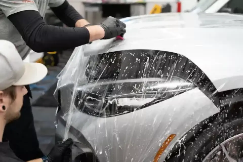 Two people apply a transparent protective film to the hood and headlight of a car.