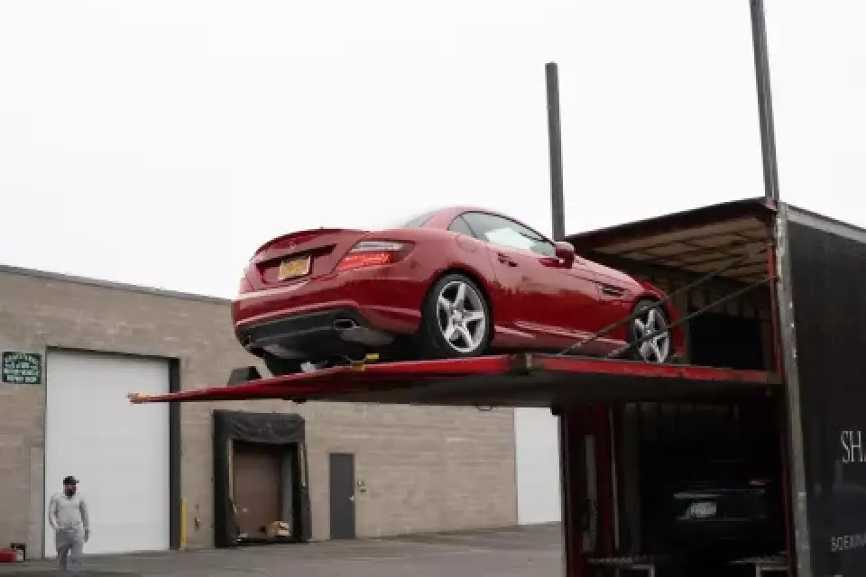 A red convertible car being loaded onto an auto transport trailer outside a warehouse.