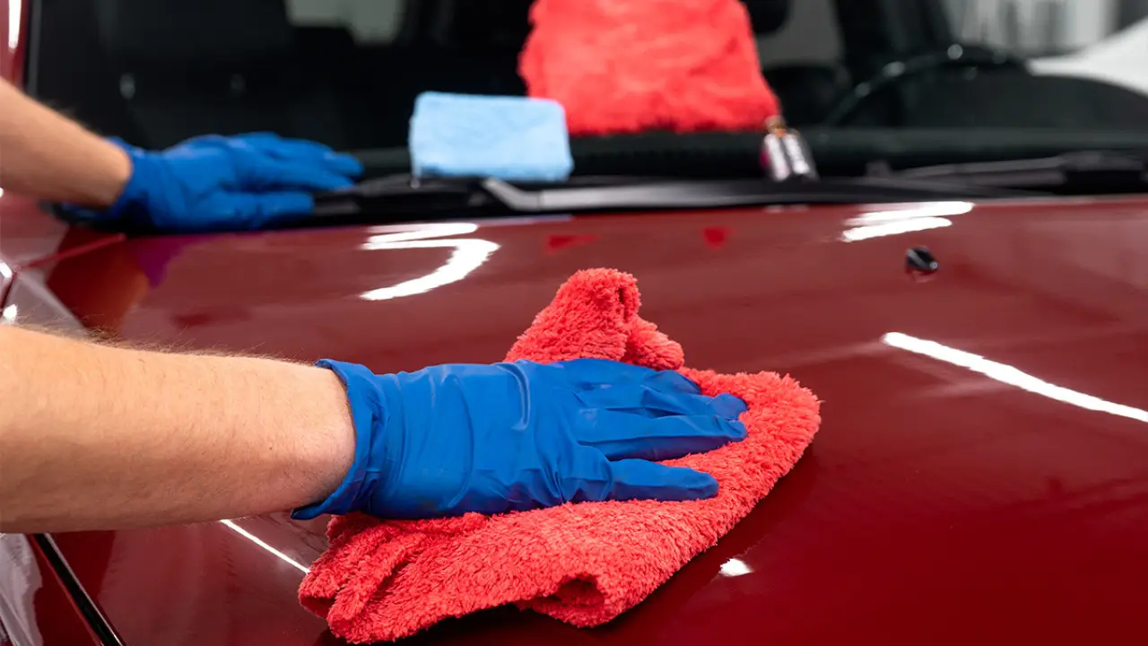 Person wearing blue gloves cleaning a red car hood with a red microfiber cloth.