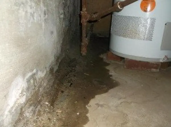 Water leaking on a basement floor near a wall and a water heater with rusted pipes above.