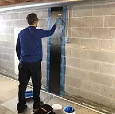 Person in blue shirt applies material to a narrow strip on a basement cinder block wall.