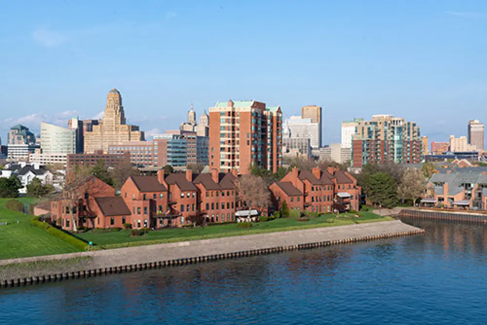 Red-brick townhouses by the water with a city skyline and blue sky in the background.