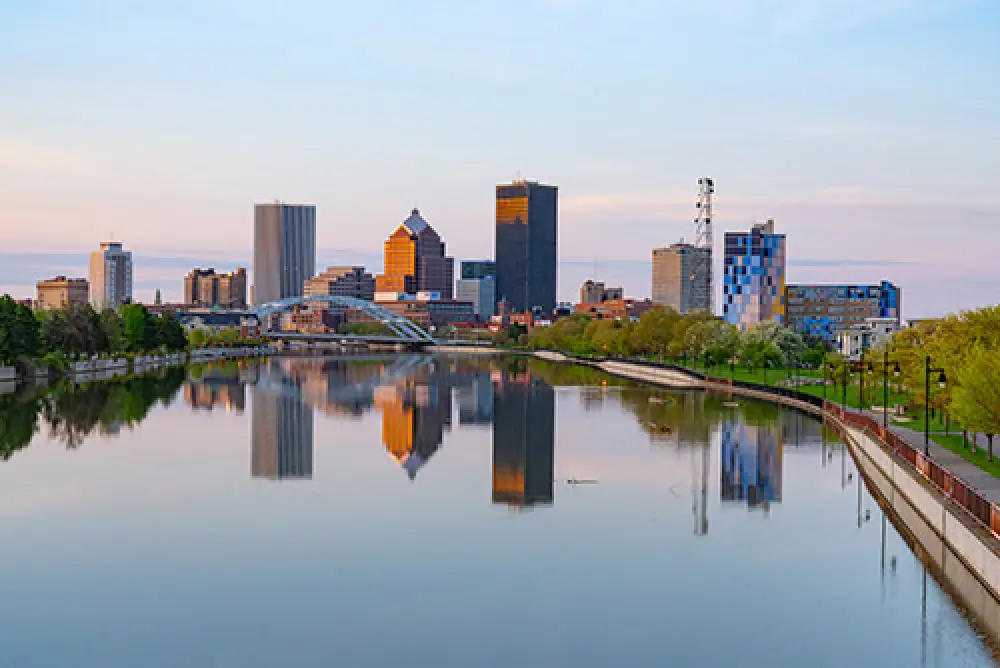 Downtown city skyline reflected in a calm river at sunset, with trees lining the riverbanks.