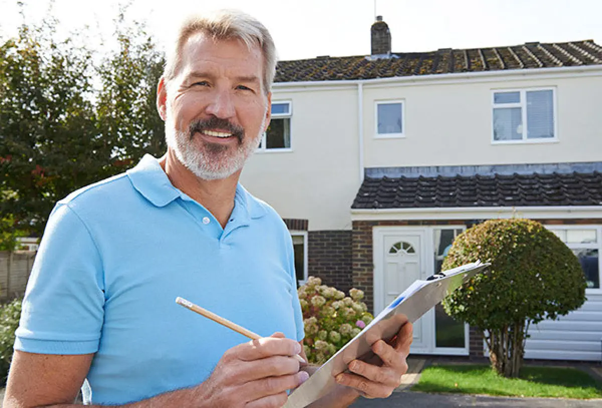 Smiling man holding clipboard and pencil, standing in front of a house on a sunny day.