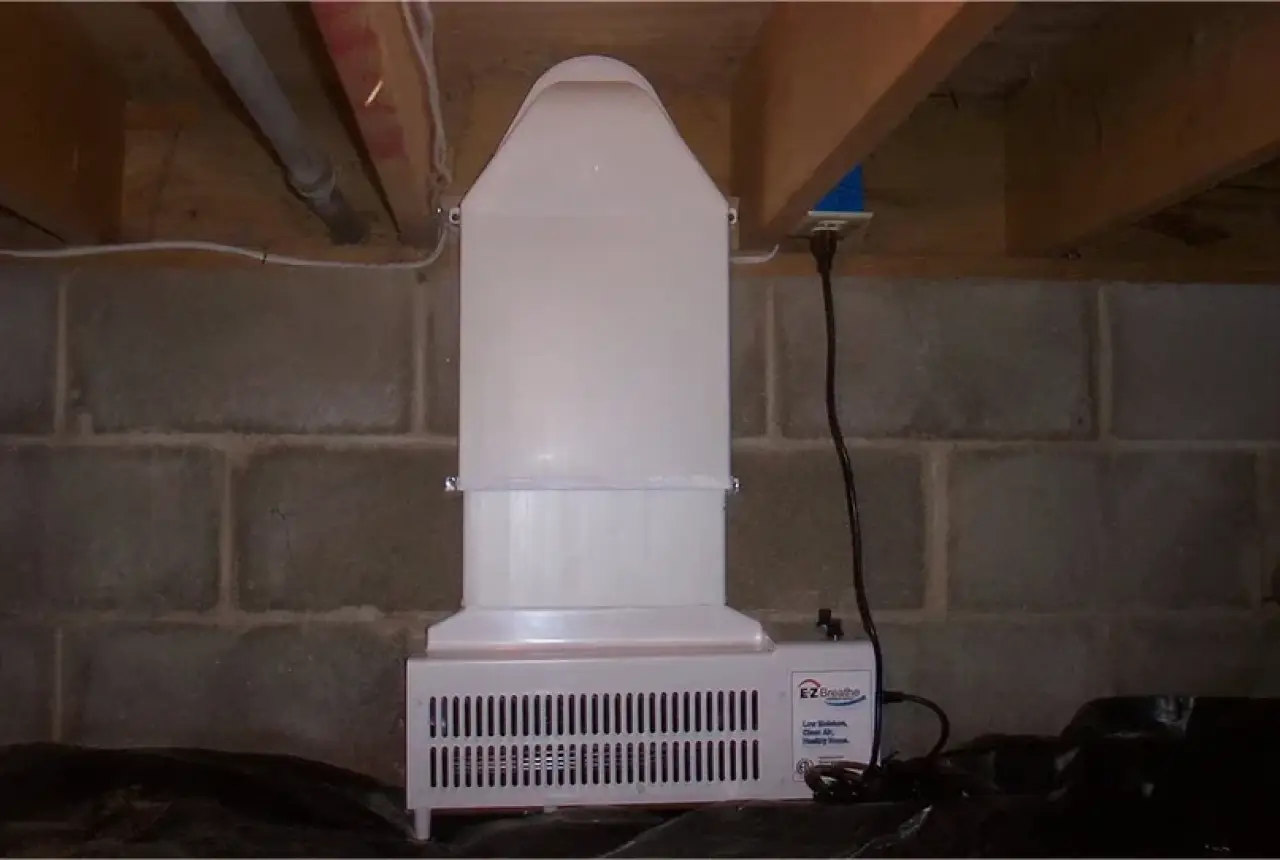 A white crawl space dehumidifier sits against a concrete block wall under exposed floor joists.