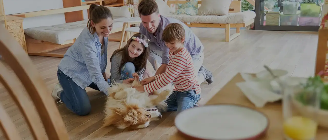 A family of four smiles and plays with their dog on the floor of a bright living room.