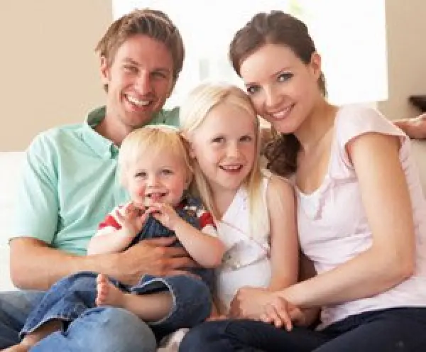 Smiling family of four, two adults and two children, sitting together on a couch indoors.