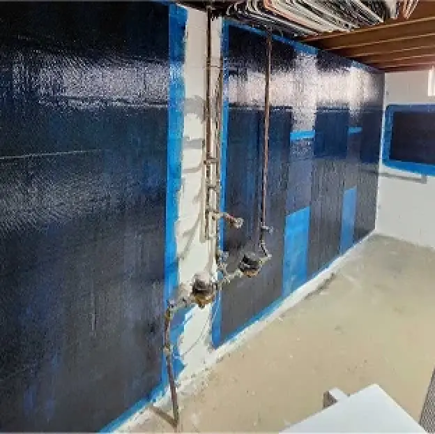 Basement wall with black waterproofing coating, exposed pipes, and partially unfinished floor.