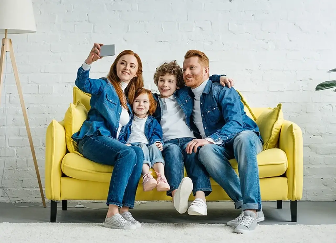 Family of four in denim outfits taking a selfie together on a yellow couch in a bright room.