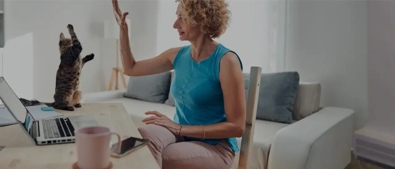 Woman giving a high five to a cat while sitting at a desk with a laptop and coffee mug.
