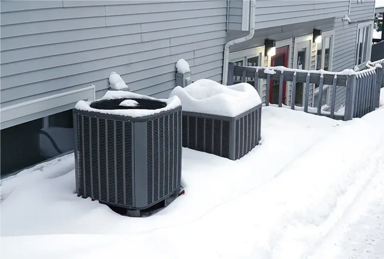 Two outdoor air conditioning units covered in snow next to a house with gray siding.