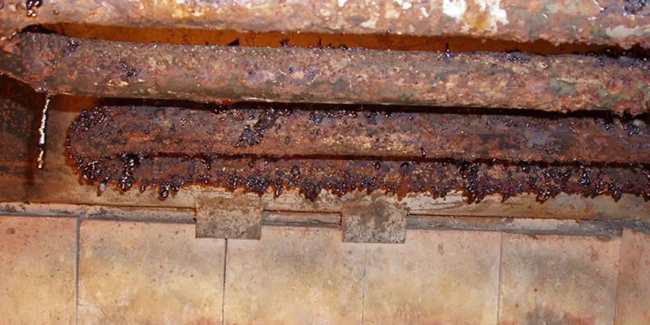Three heavily corroded metal pipes with rust and grime, above discolored tiled surfaces.