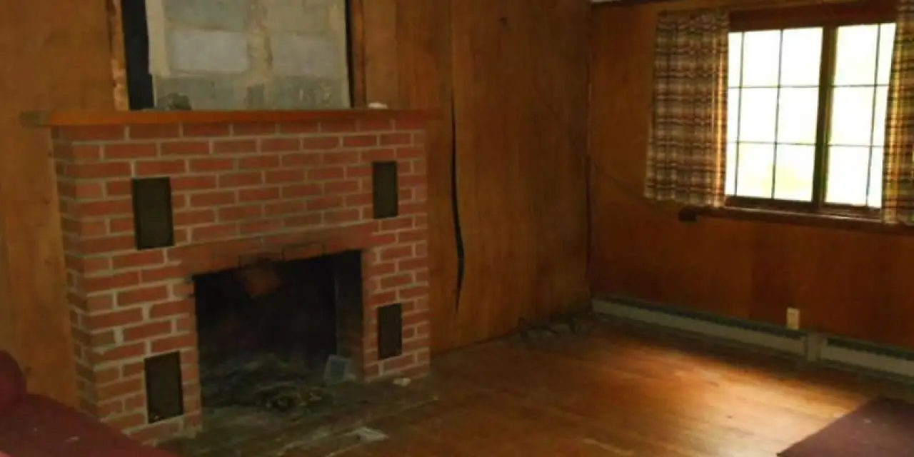 An empty room with wood-paneled walls, a brick fireplace, and a window with plaid curtains.