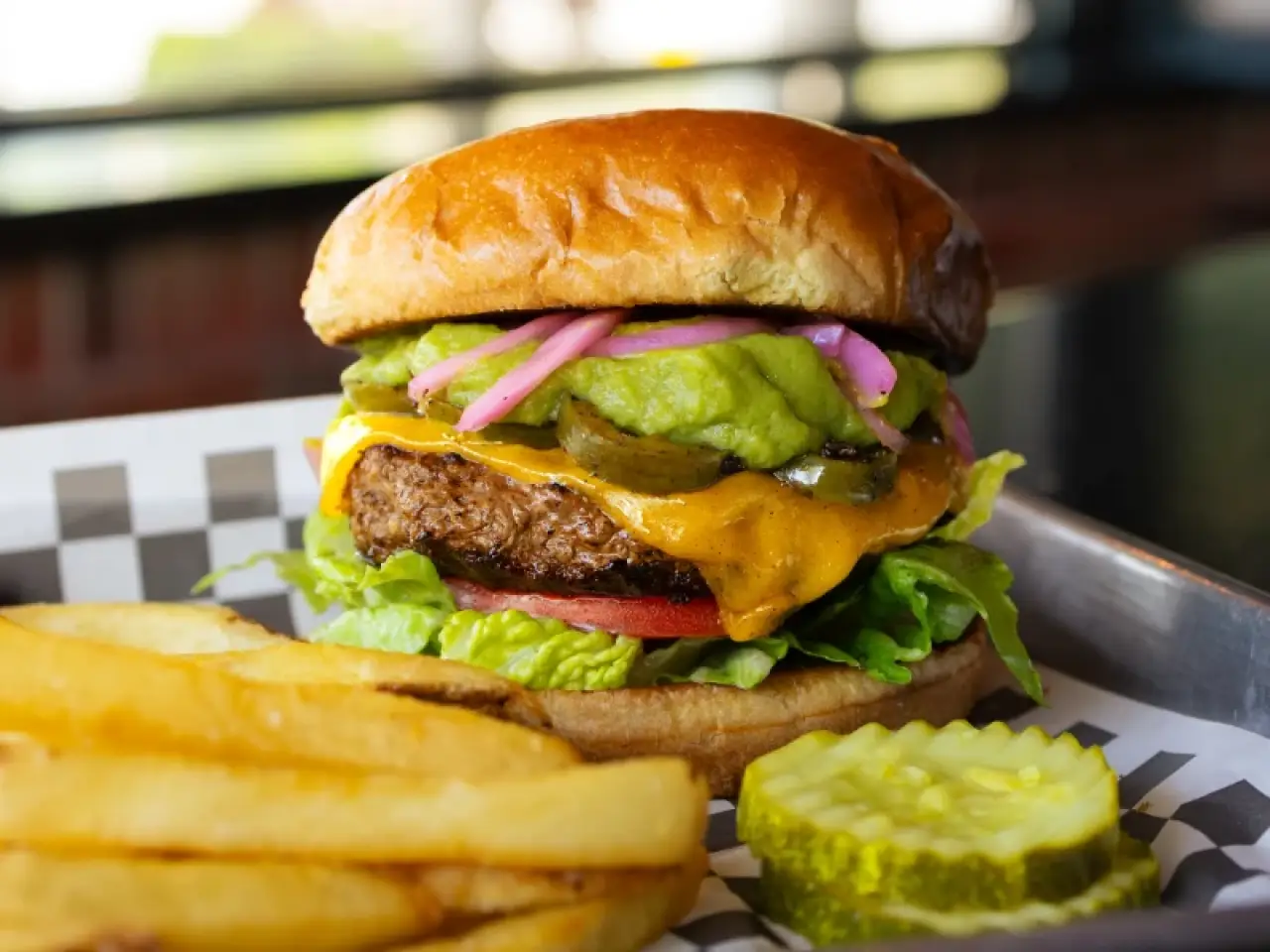 A cheeseburger with fries and pickles, topped with guacamole, onions, and lettuce.