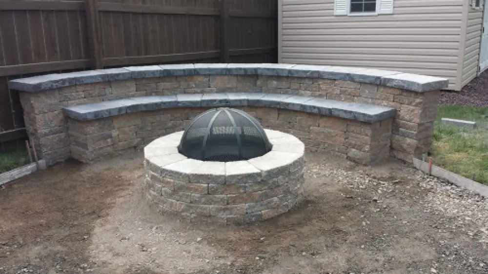 A stone fire pit with a mesh cover and curved stone bench seating in a backyard.
