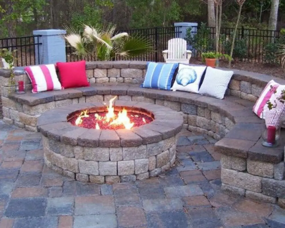 Stone fire pit with curved bench seating, colorful pillows, and a paved patio in a garden.