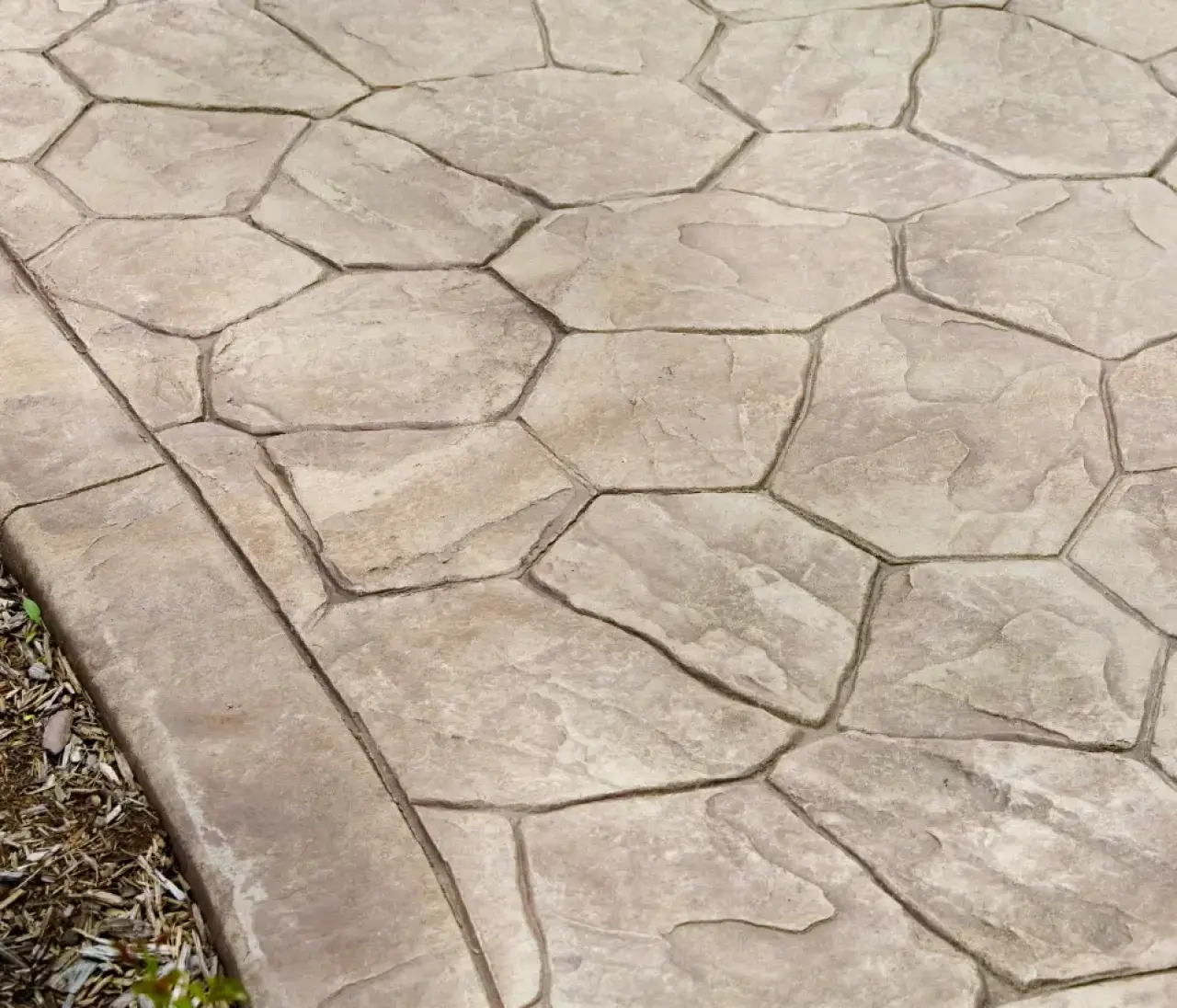 Tan stamped concrete pavement with irregular hexagonal patterns bordered by soil and mulch.